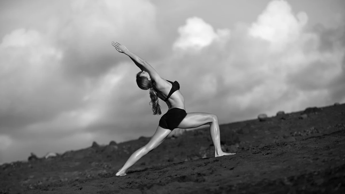 Eine Frau in der Yogastellung des Kriegers auf einem Vulkanberg aus schwarzem Sand mit einer großen Anzahl von Wolken am Himmel, die eine mystische Atmosphäre schaffen.
