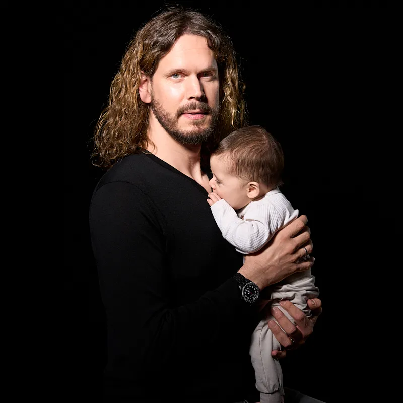 Porträt eines Mannes mit langem Haar, der ein Baby schützend im Arm hält, vor dunklem Hintergrund